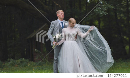 Newlywed couple posing for wedding photos in a park, holding hands and a bouquet, with the bride's veil flowing in the wind 124277011