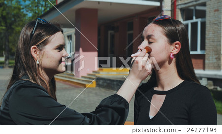 Two high school girls using a makeup brush during a school break, having fun together Two high school girls using a makeup brush during a school break, having fun together 124277193