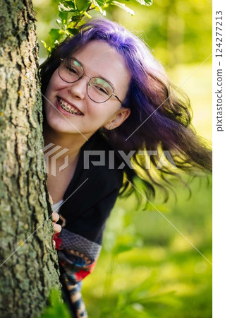 Teenage girl with purple hair, glasses, and braces peeking from behind tree bark during bright summer day Teenage girl with purple hair, glasses, and braces peeking from behind tree bark during bright summer day 124277213