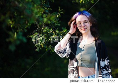 Teenager with purple hair and glasses enjoying a peaceful moment in nature, touching her hair under the shade of a tree Teenager with purple hair and glasses enjoying a peaceful moment in nature, touching her hair under the shade of a tree 124277222