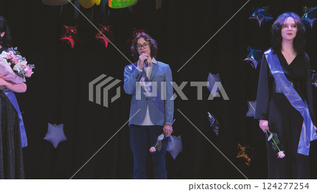 Schoolboy wearing a sash giving a speech during his graduation ceremony with his friends, holding a microphone and a rose Schoolboy wearing a sash giving a speech during his graduation ceremony with his friends, holding a microphone and a rose 124277254