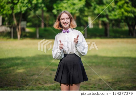 Excited student in a white shirt and bow tie giving a thumbs up on their first day of school in a park 124277379