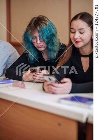 Two female students are using their smartphones during a class at university or college, potentially engaging in social media or other online activities 124277381