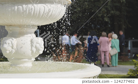 Pristine white fountain spraying water droplets, blurred wedding celebration unfolding behind, radiating romantic elegant ambiance Pristine white fountain spraying water droplets, blurred wedding celebration unfolding behind, radiating romantic elegant ambiance 124277402