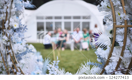 Wedding guests gathered, socializing near white tent, framed by lush floral arch, celebrating nuptial moment outdoors Wedding guests gathered, socializing near white tent, framed by lush floral arch, celebrating nuptial moment outdoors 124277413