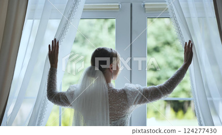 White gowned bride pulling back curtains, gazing through window at sunlit garden inside elegant hotel room White gowned bride pulling back curtains, gazing through window at sunlit garden inside elegant hotel room 124277430