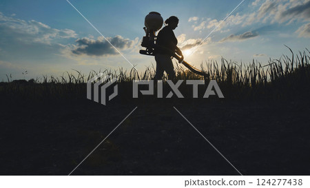 Farmer walking on cultivated field spraying pesticides with manual knapsack sprayer at sunset 124277438