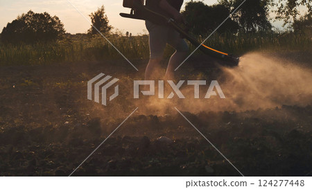 Farmer walking on cultivated field spraying pesticides with manual knapsack sprayer at sunset 124277448