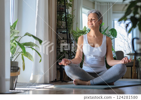 Beautiful smiling gray-haired mature middle-aged woman at home sitting on yoga mat in lotus position Beautiful smiling gray-haired mature middle-aged woman at home sitting on yoga mat in lotus position 124281580