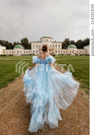 Woman in light blue ball gown runs along a path against the backdrop of a historic estate.Back view. Woman in light blue ball gown runs along a path against the backdrop of a historic estate.Back view. 124281659