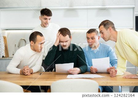 Friends reading financial documents and trying to solve problems together while sitting at table in their kitchen 124282164