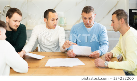Friends reading financial documents and trying to solve problems together while sitting at table in their kitchen 124282200