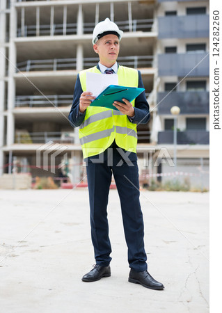 Architect in protective helmet and jacket with folder of documents on construction site 124282260