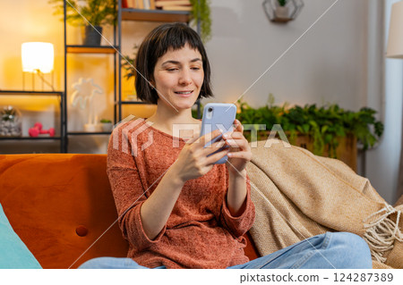 Happy woman thinking and text messaging on smartphone watching online movie sitting on home sofa 124287389