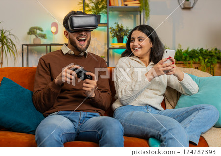Happy young man wearing VR goggles playing videogame with joystick sitting beside Indian girlfriend 124287393