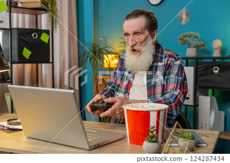 Excited happy senior man celebrating victory in play video game on laptop using controller joystick 124287434