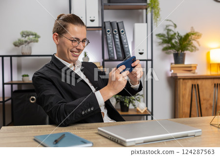 Excited young businessman playing shooter or racing video games on smartphone sitting at desk 124287563