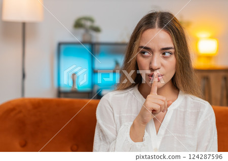 Portrait of young woman on sofa presses index finger to lips makes silence hush gesture sign at home Portrait of young woman on sofa presses index finger to lips makes silence hush gesture sign at home 124287596