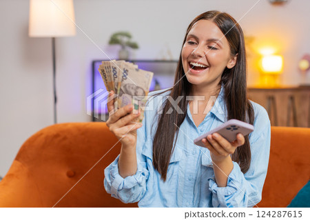 Smiling happy rich young woman counting money hryvnias cash, use smartphone income saves lottery win 124287615
