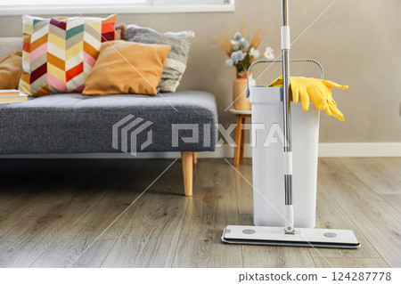 Cleaning supplies: mop, gloves, bucket with wringer sitting on wooden laminate floor in modern living room, wet equipment tools, housekeeping concept. Emphasizing home sanitation, cleanliness, hygiene 124287778