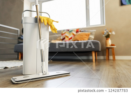 Cleaning supplies: mop, gloves, bucket with wringer sitting on wooden laminate floor in modern living room, wet equipment tools, housekeeping concept. Emphasizing home sanitation, cleanliness, hygiene 124287783