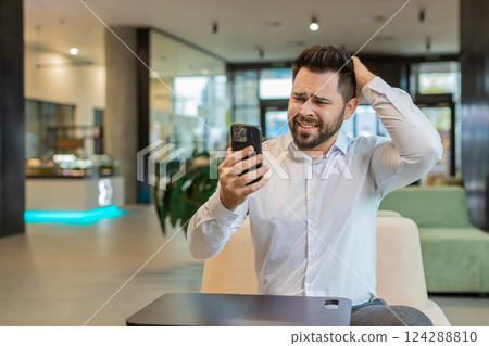 Young businessman using mobile phone becomes shocked sudden lottery results in office hotel lobby 124288810