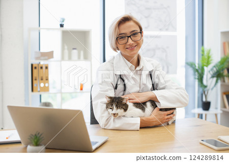 Young adult female veterinarian in white coat with stethoscope caring for domestic cat in clinic. Professional pet healthcare and animal care concept. Bright office setting with plants and equipment. Young adult female veterinarian in white coat with stethoscope caring for domestic cat in clinic. Professional pet healthcare and animal care concept. Bright office setting with plants and equipment. 124289814