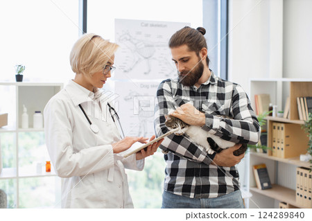 Female veterinarian discussing treatment plan with adult bearded man holding cat in clinic. Medical professional explaining pet care using tablet demonstrating attentive and professional consultation 124289840