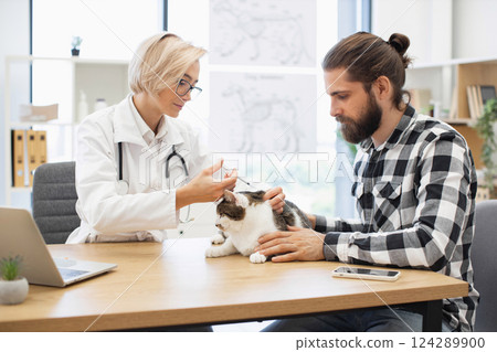 Female veterinarian administering vaccination to domestic cat, accompanied by young Caucasian adult male owner 124289900
