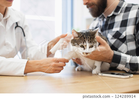 Veterinarian places splint on small cat's injured paw assisted by young adult owner. Scene shows care and tenderness in medical setting during treatment session. 124289914