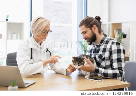 Male owner and female veterinarian discuss care of injured cat in clinic room. Veterinarian treats cat's injury, showing care and expertise, while owner shows concern and care for pet's wellbeing. 124289916
