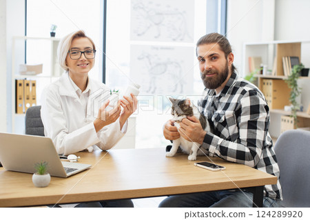 Male pet owner holding cat while visiting female veterinarian discussing medicine and pet care. Professional veterinary guidance for pet health. Middle-aged man, middle-aged woman healthcare provider. Male pet owner holding cat while visiting female veterinarian discussing medicine and pet care. Professional veterinary guidance for pet health. Middle-aged man, middle-aged woman healthcare provider. 124289920