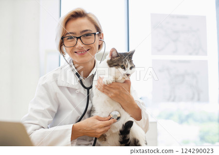 Female veterinarian wearing glasses examining a tabby cat with a stethoscope in clinic. Scene involves a young blonde adult focused on ensuring proper care. 124290023