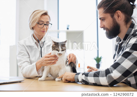 Veterinarian uses stethoscope to examine cat while owner assists. Scene involves adult Caucasian male and female interacting with pleasant atmosphere in pet healthcare environment. Veterinarian uses stethoscope to examine cat while owner assists. Scene involves adult Caucasian male and female interacting with pleasant atmosphere in pet healthcare environment. 124290072