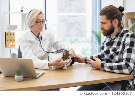 Female veterinarian using stethoscope on domestic cat held by bearded young adult man. Modern veterinary clinic with laptop and anatomical posters visible. 124290107