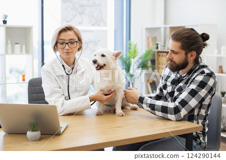 Professional female veterinarian checks healthy white dog at modern clinic with young male owner observing. Scene emphasizes care, trust, animal health. Models include young adult male, young female. 124290144