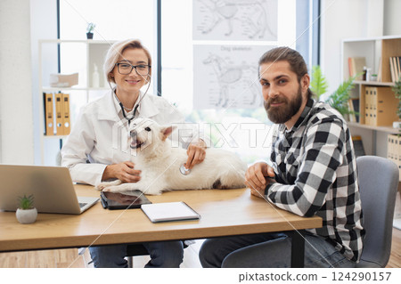 Female veterinarian examining small white dog with stethoscope, owner sitting nearby. Office setting includes medical posters. Represents veterinary care, pet companionship, professional consultation Female veterinarian examining small white dog with stethoscope, owner sitting nearby. Office setting includes medical posters. Represents veterinary care, pet companionship, professional consultation 124290157