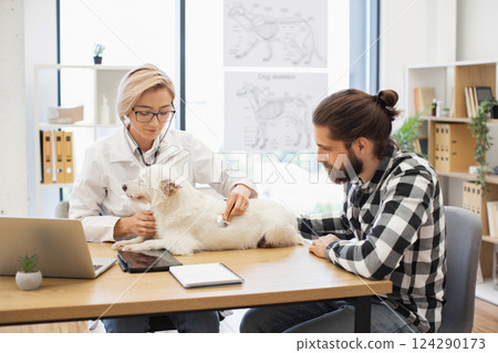 Female veterinarian conducting health examination of a dog while male owner observes session. Scene depicts professional veterinary care, showcasing careful pet owner interaction in clinic. 124290173