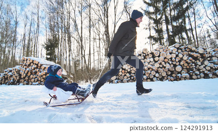 Winter scene featuring smiling father pulling laughing child on wooden sled through snow covered forest near wood pile Winter scene featuring smiling father pulling laughing child on wooden sled through snow covered forest near wood pile 124291913