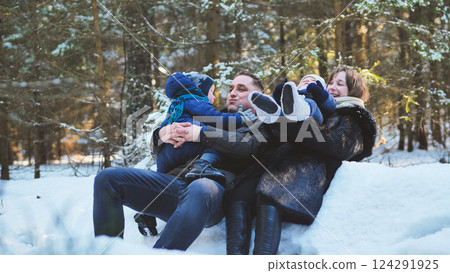 Parents holding their children while sitting on snow in a winter forest, enjoying a sunny winter day Parents holding their children while sitting on snow in a winter forest, enjoying a sunny winter day 124291925
