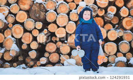 Cheerful kid bundled up, holding mugs and mandarins, standing amid snowy landscape with wooden logs, radiating winter joy 124291931