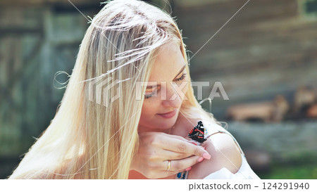 Young blond woman holding a colorful butterfly on her bare shoulder, enjoying a sunny summer day outdoors 124291940