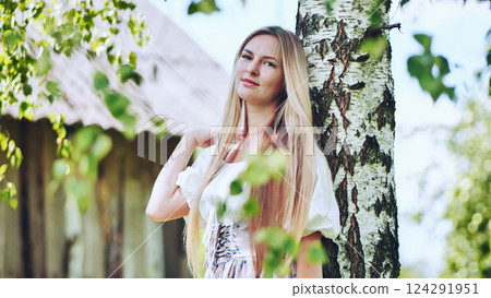 Young blonde woman with long hair wearing traditional slavic dress posing next to birch tree in a rural setting 124291951