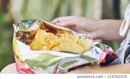 Woman relaxing outdoors, munching crispy potato chips, savoring salty treat during bright afternoon sunlight Woman relaxing outdoors, munching crispy potato chips, savoring salty treat during bright afternoon sunlight 124292019