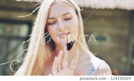 Blonde woman gently holding red admiral butterfly on finger, radiating serene connection with vibrant summer landscape 124292024