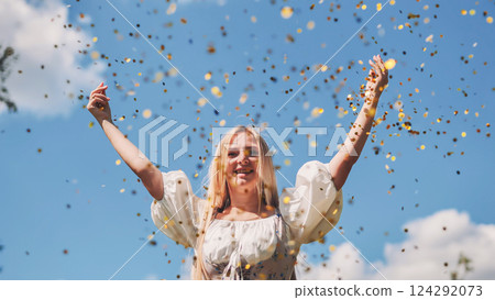 Young blonde woman throwing gold confetti in the air, celebrating a joyful event under a sunny blue sky Young blonde woman throwing gold confetti in the air, celebrating a joyful event under a sunny blue sky 124292073