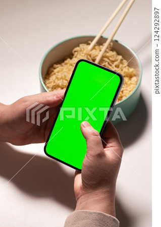 Female using smartphone while eating pasta at home. Smartphone with green screen with space for text. Smartphone in hands with served table in background 124292897