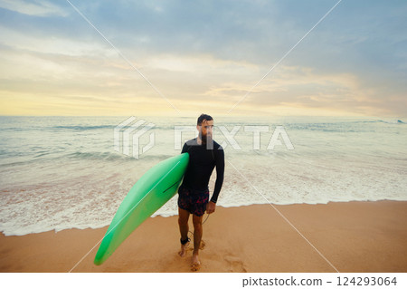 Smiling young man with surfboard on the beach. Boy coming out of the ocean after water surfing. 124293064