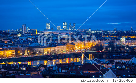 Historic National Theatre stands illuminated beside modern Pankrac buildings. The Prague skyline sparkles at dusk, reflecting along the Vltava River's surface. 124293409