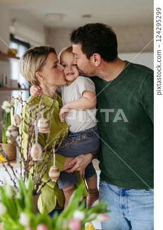 Parents standing next to pussy willows branches with easter eggs on them, kissing little boy on cheeks. 124295299